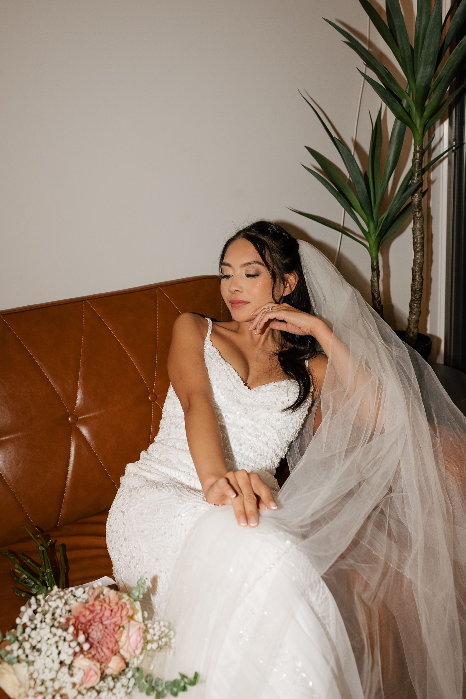 A bride in a white lace wedding dress sits gracefully on a brown leather couch with flowers and greenery nearby.