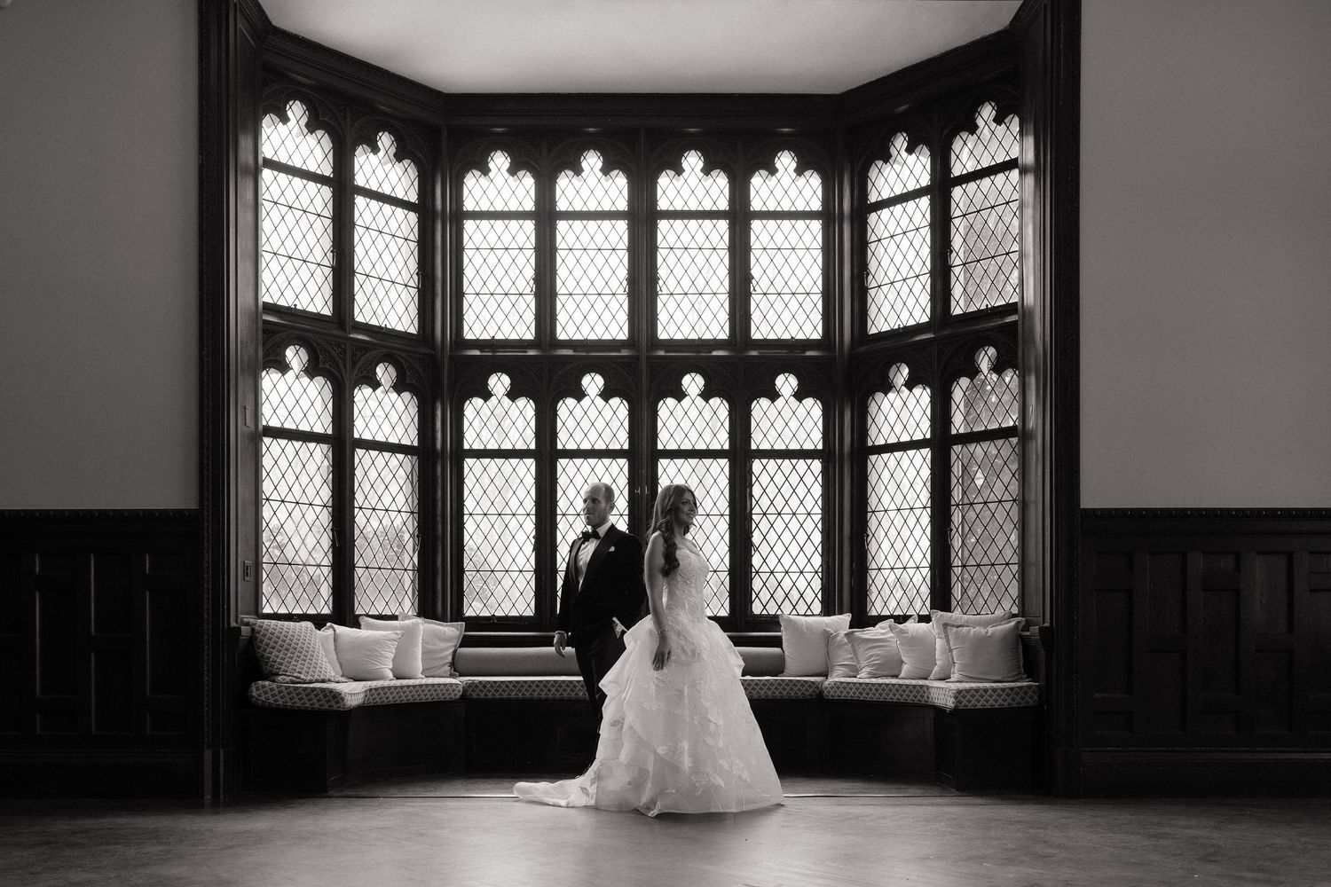 Black and white wedding photo of bride and groom silhouetted before gothic arched windows in elegant venue.