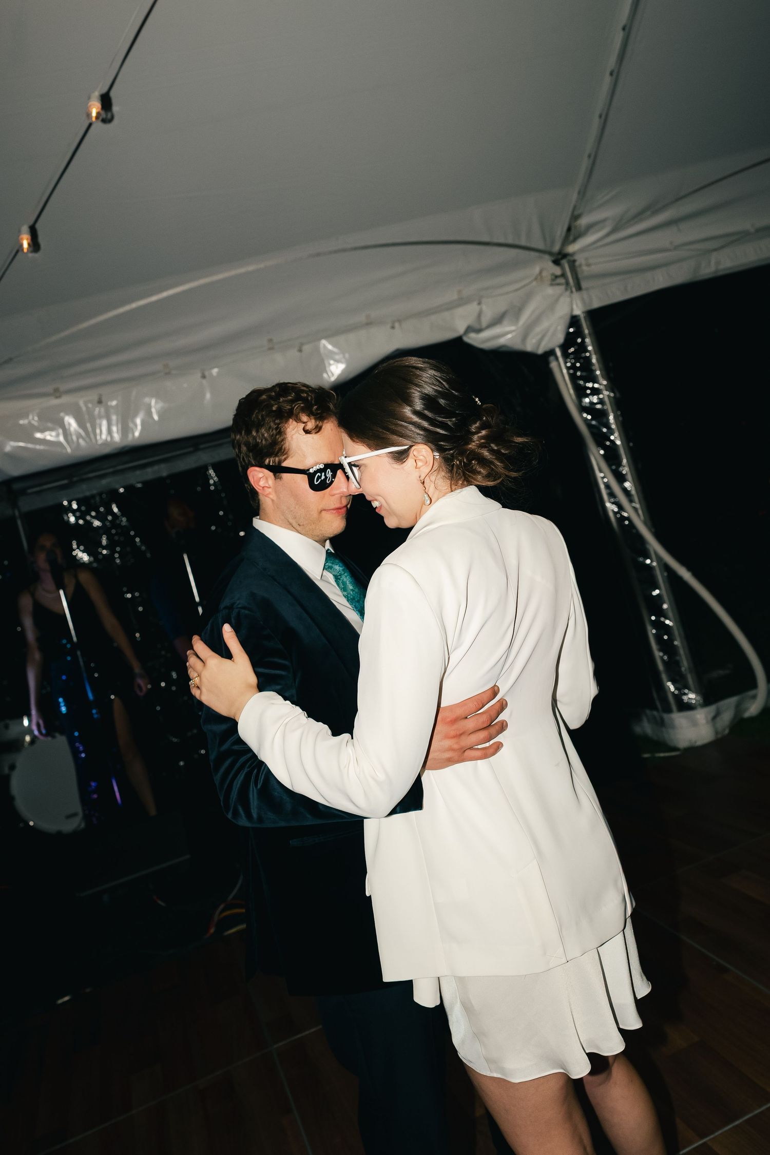 Couple shares first dance under white tent at wedding reception, groom in navy suit with teal tie and sunglasses.