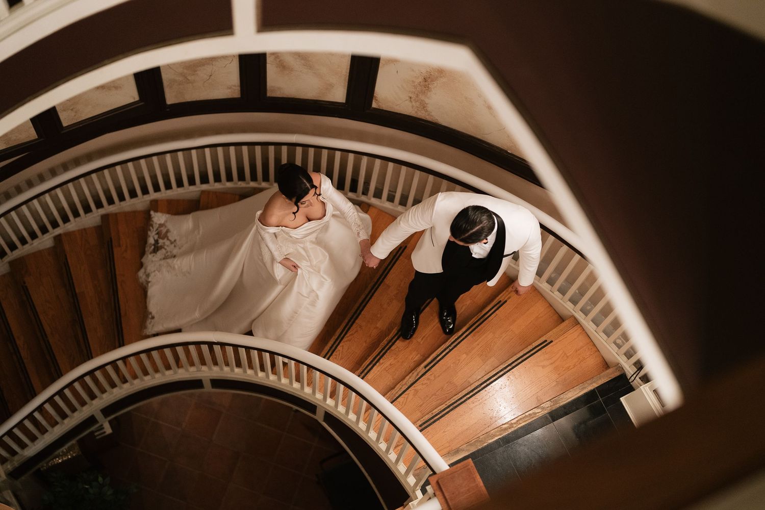 A spiral staircase viewed from above shows a wedding couple descending the elegant curved wooden steps.