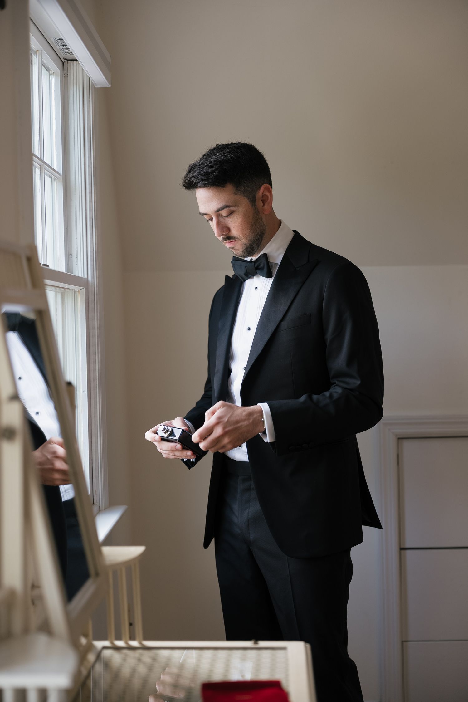 A figure in a black tuxedo stands by a window while looking down at a mobile device in a sunlit room.