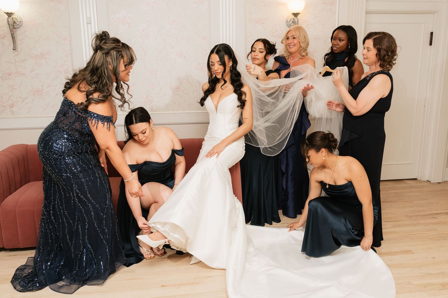 Bridesmaids helping bride into wedding dress and veil in elegant white dressing room before ceremony.