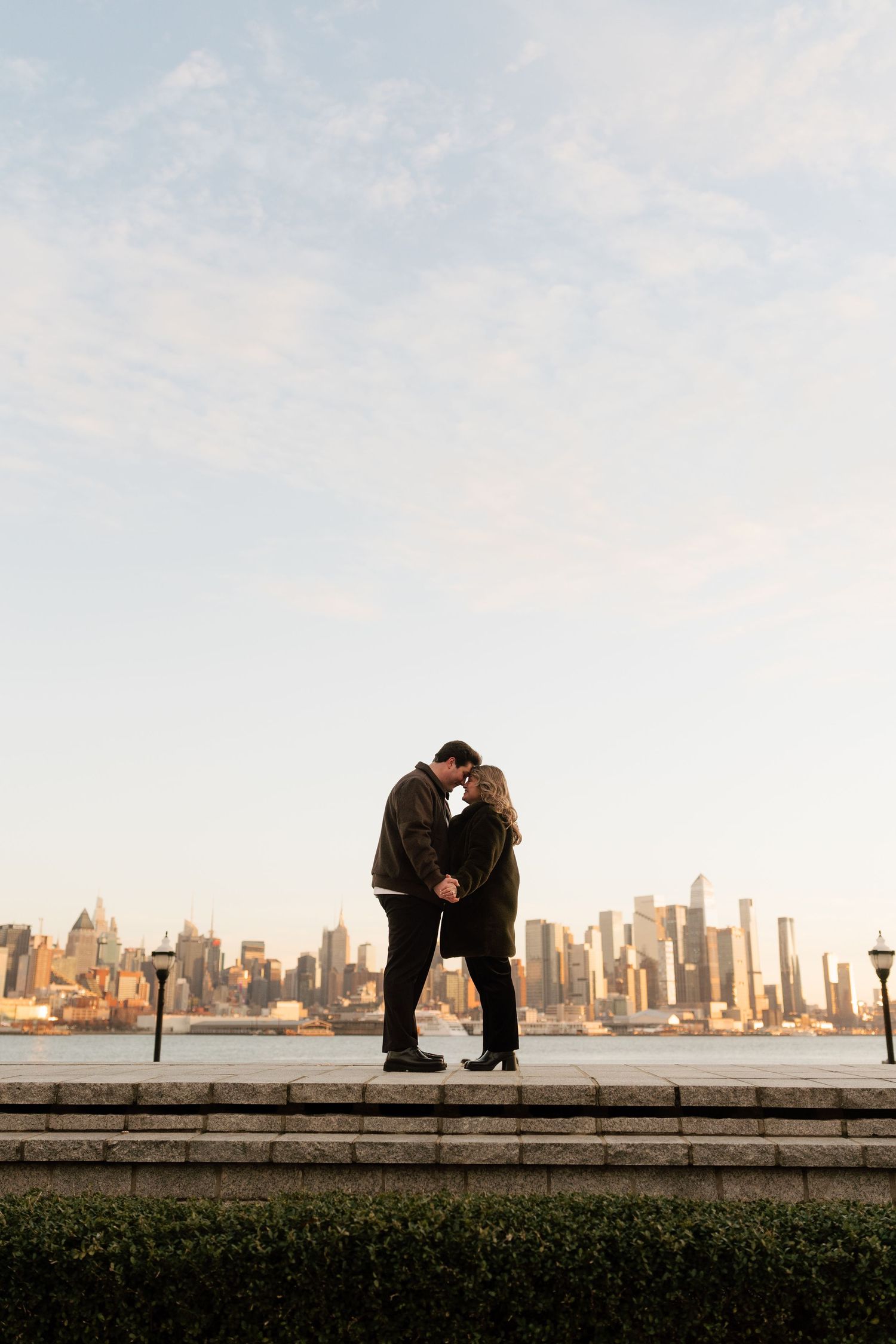 Couple sharing a romantic kiss on steps with the New York City skyline and Hudson River at golden hour.
