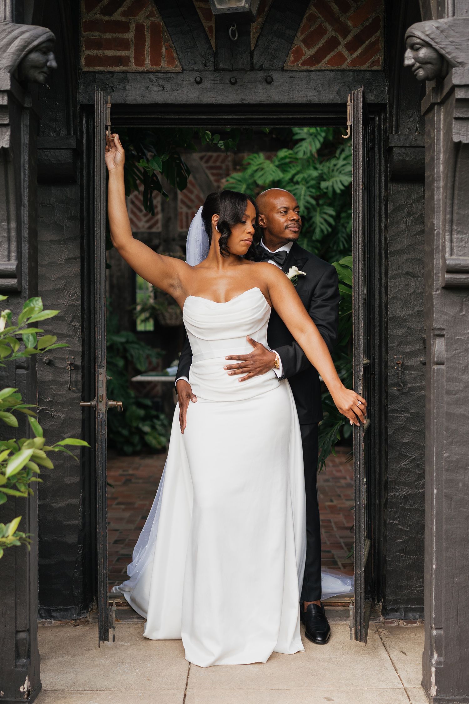 Bride in strapless white gown and groom in tuxedo pose elegantly in ornate dark doorway.
