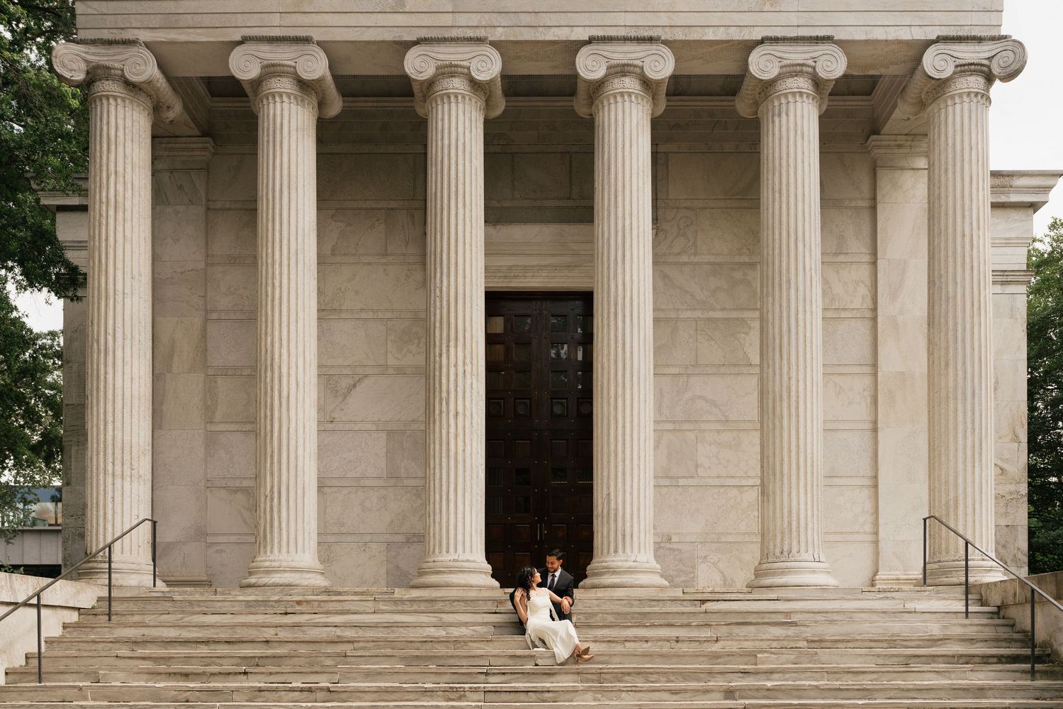 Couple sitting on steps of a neoclassical building with tall Ionic columns and grand stone architecture.