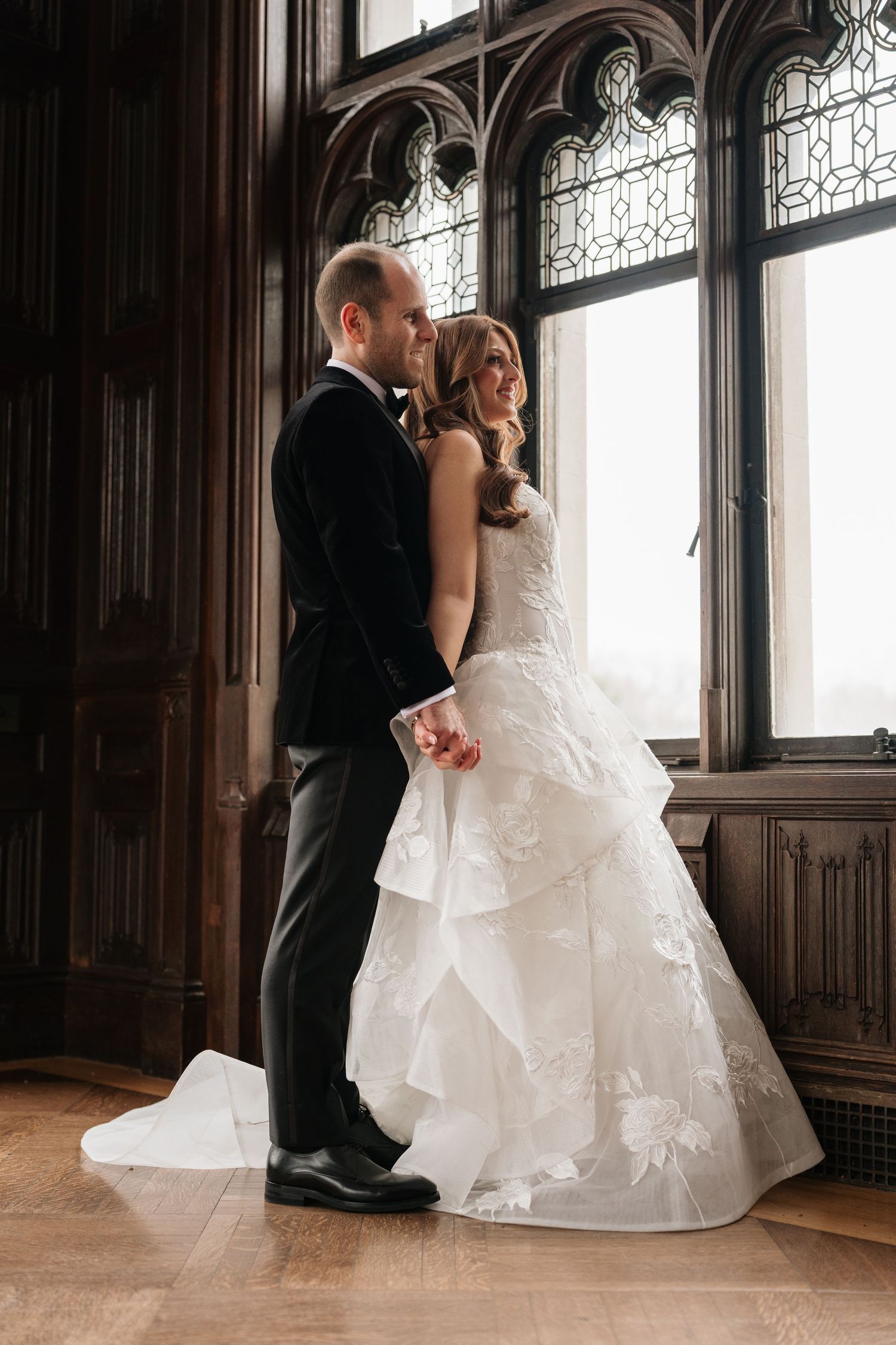 Bride in layered white gown and groom in black tuxedo holding hands by ornate Gothic window in elegant venue.