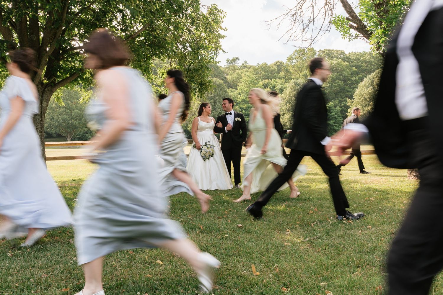 Wedding party running across green lawn with bride and groom standing still in center, outdoor summer ceremony.