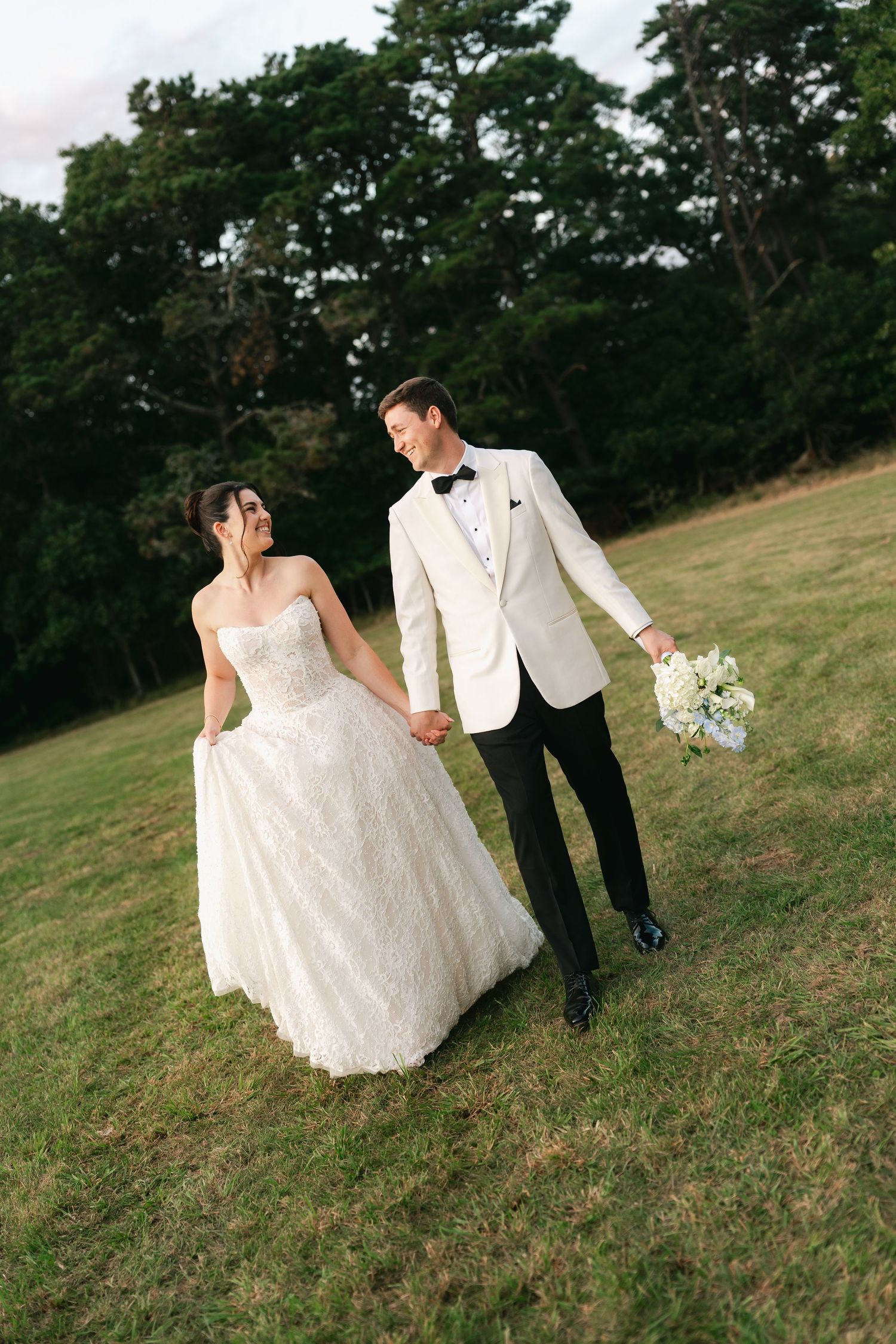 Bride in white gown and groom in ivory tuxedo hold hands walking across green lawn with trees behind.