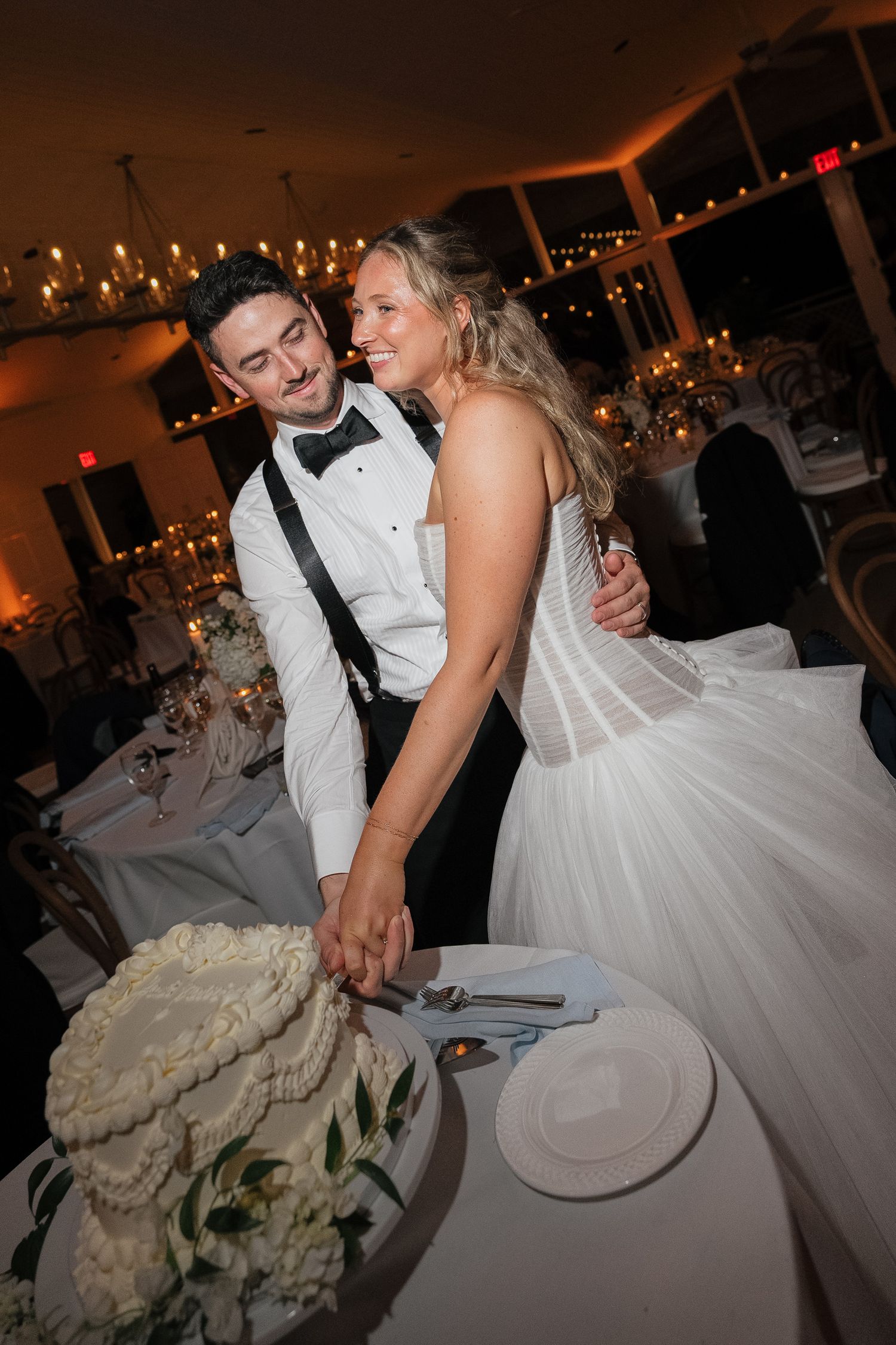 A couple in formal wedding attire shares an intimate moment while cutting their wedding cake at a reception.