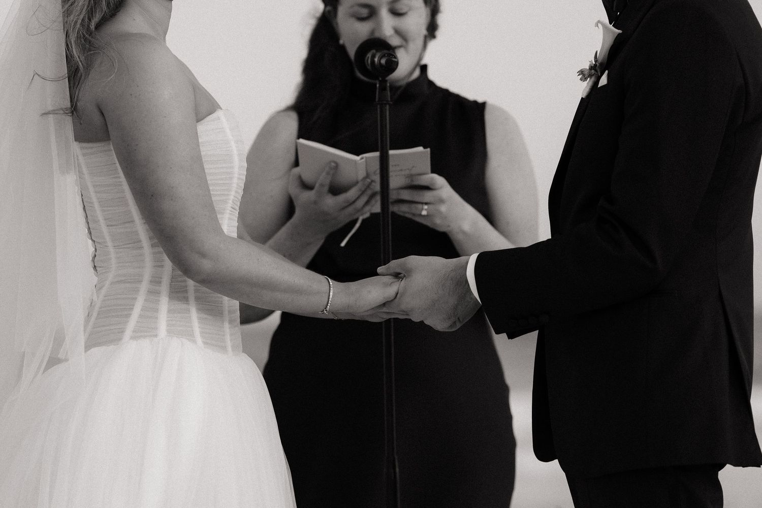 Black and white photo of a wedding ceremony with couple holding hands as officiant reads from book during service.