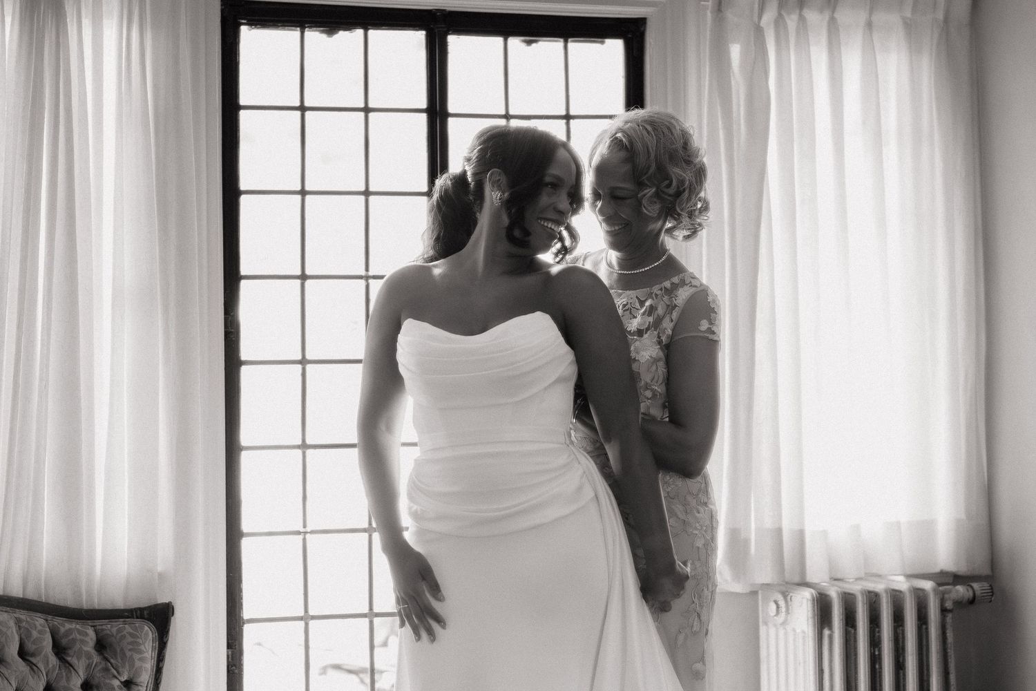 A black and white photograph of two people in formal attire standing by a window with white curtains.