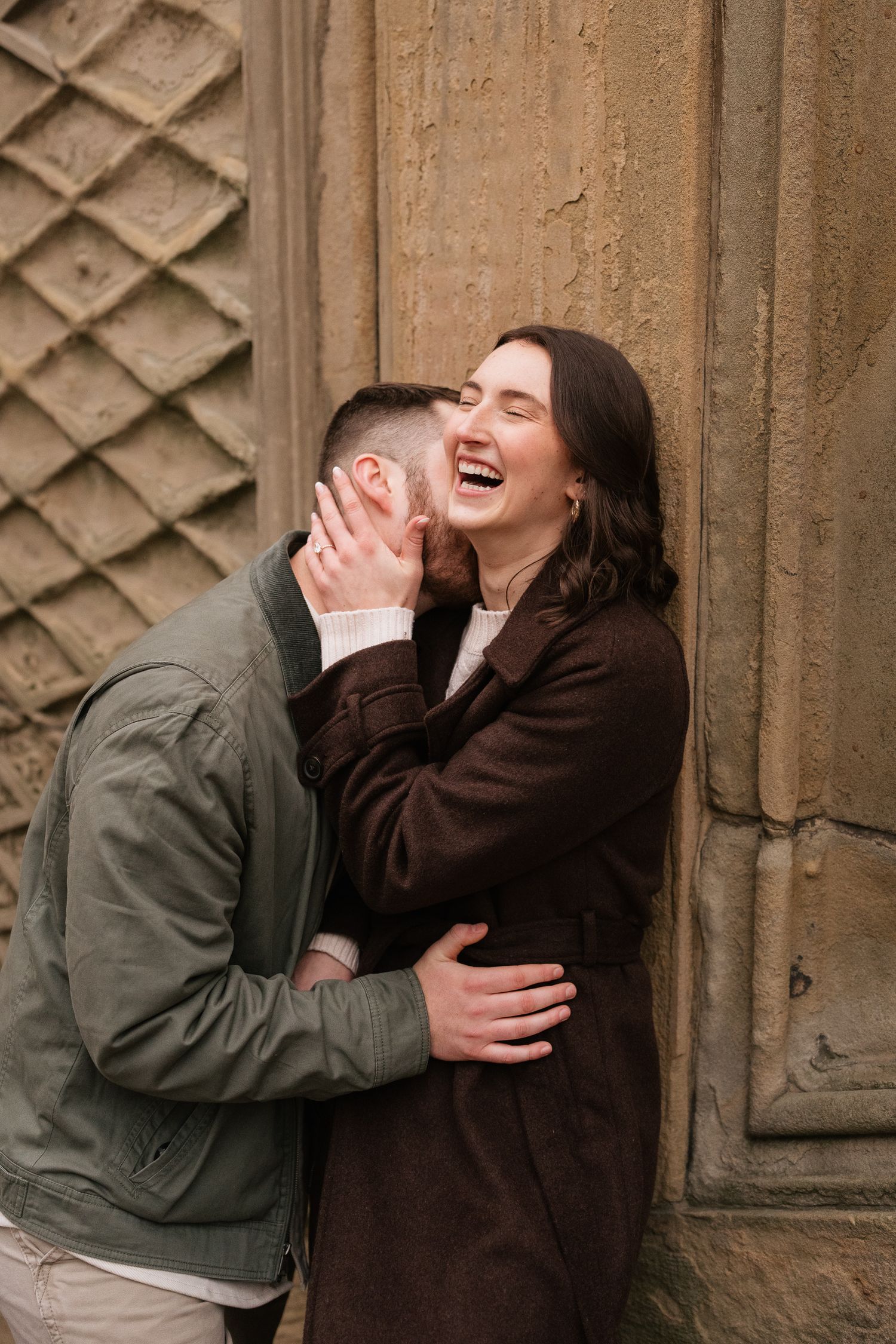 A joyful couple embracing and laughing together against an ornate stone wall, wearing casual autumn jackets.