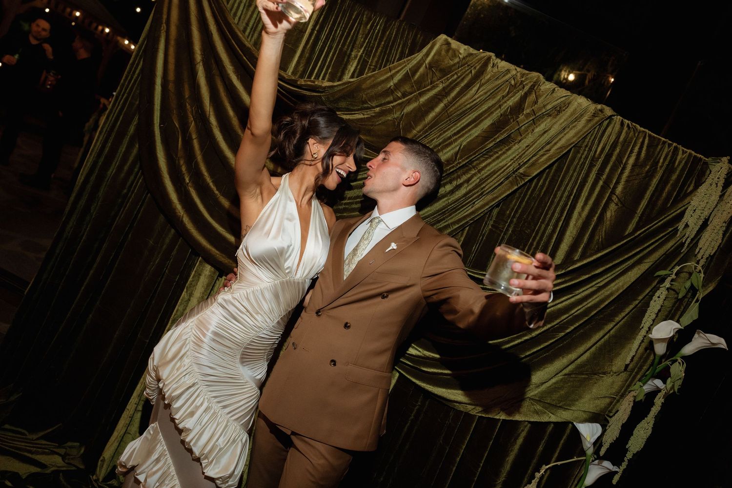 Bride in white gown and groom in tan suit dancing joyfully in front of green velvet curtain backdrop.