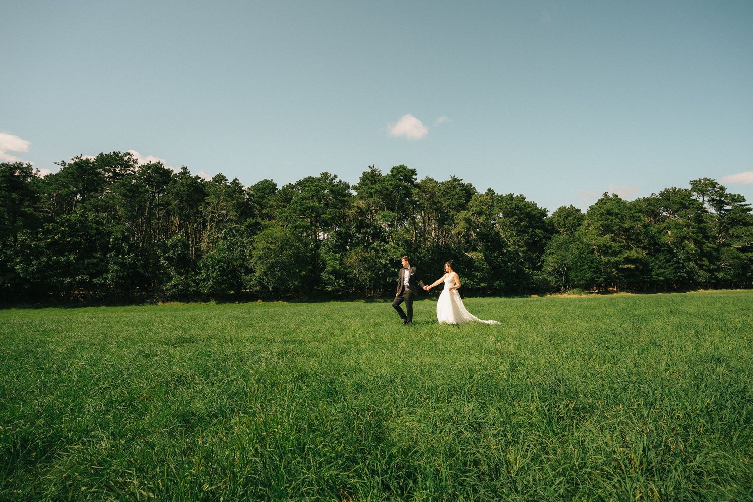 A bride and groom walk hand in hand across a lush green field with a forest backdrop on their wedding day.