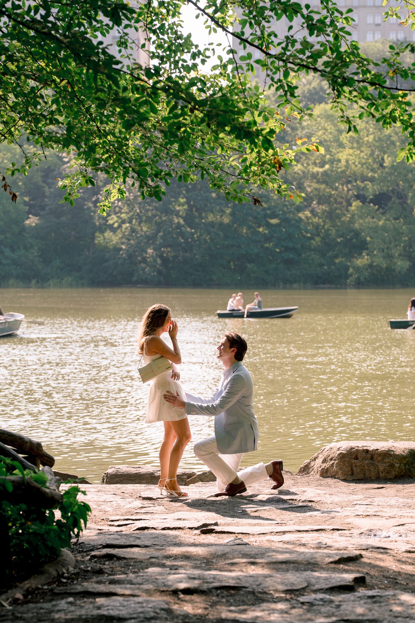 A romantic lakeside proposal scene unfolds on a rocky shore under overhanging tree branches at sunset.