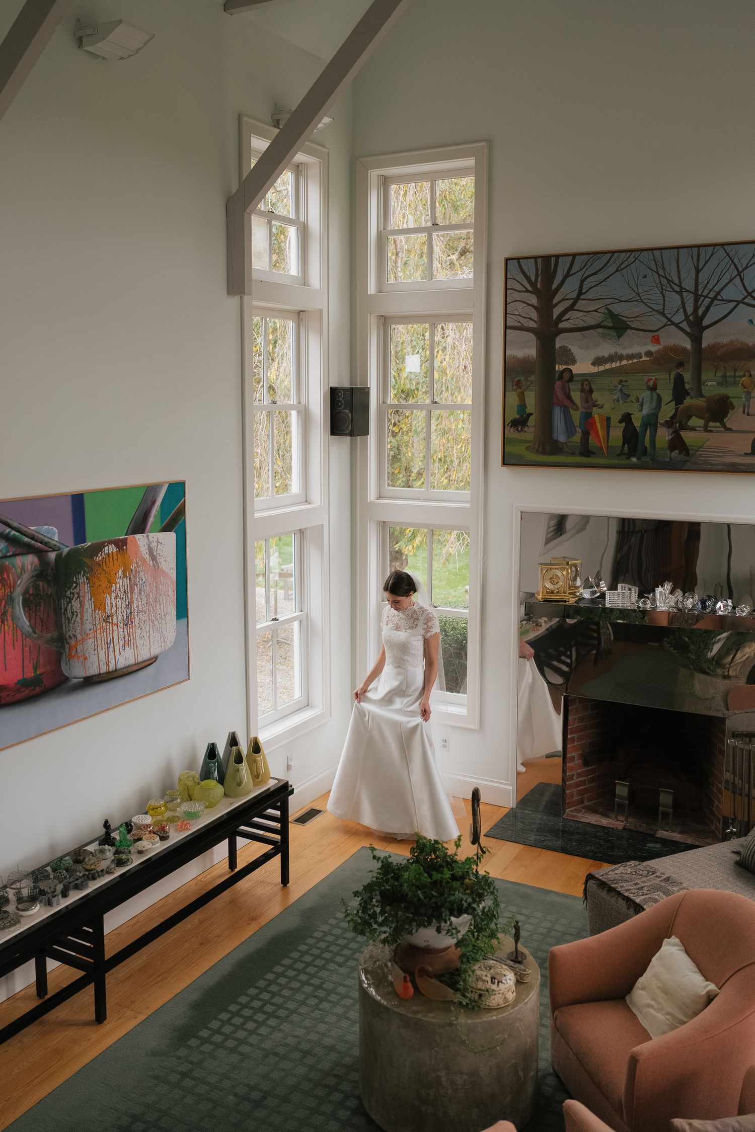 Bride in white gown stands by tall windows in elegant living room with fireplace and colorful artwork.