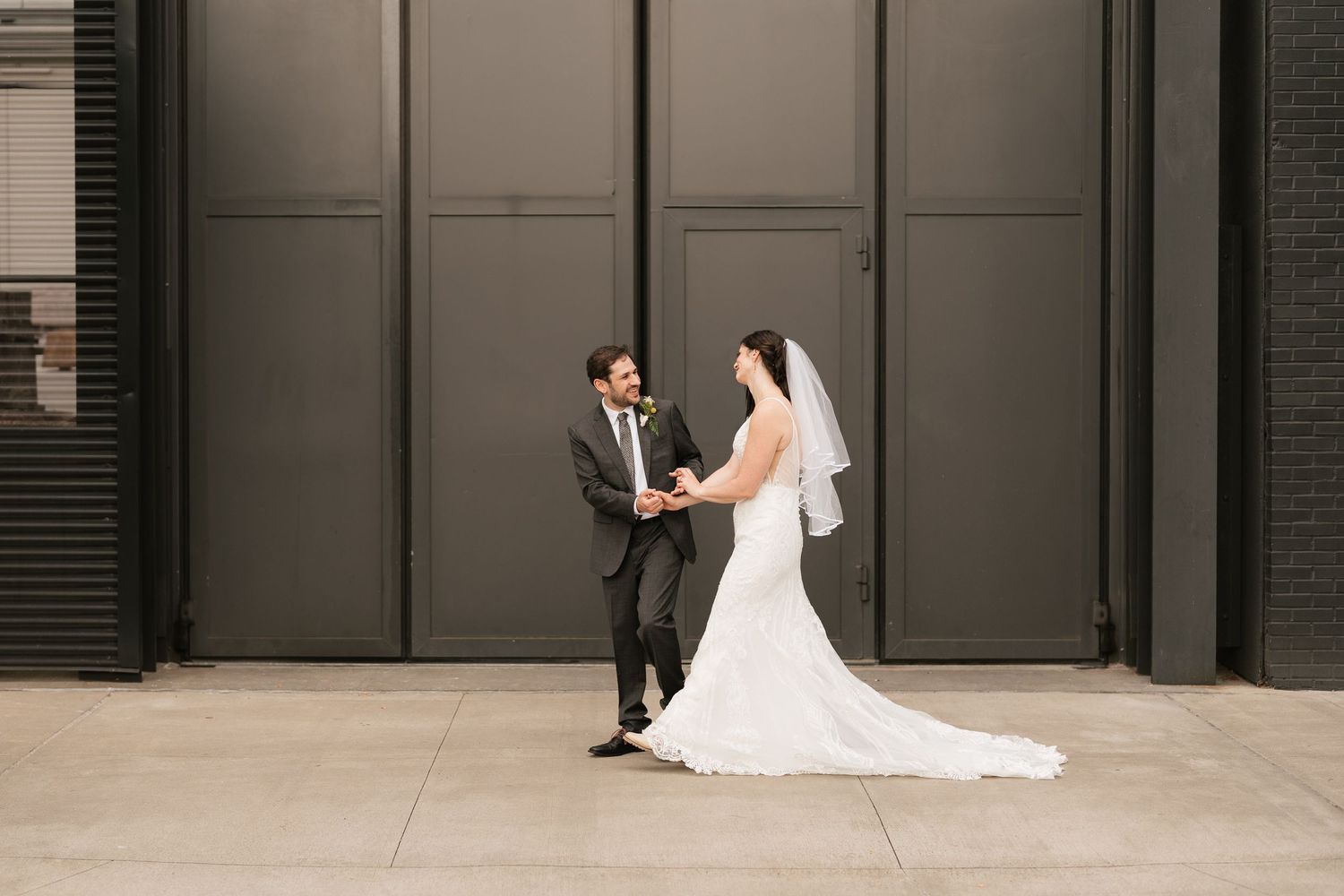 Bride and groom hold hands facing each other in front of a dark gray wall during outdoor wedding portraits.