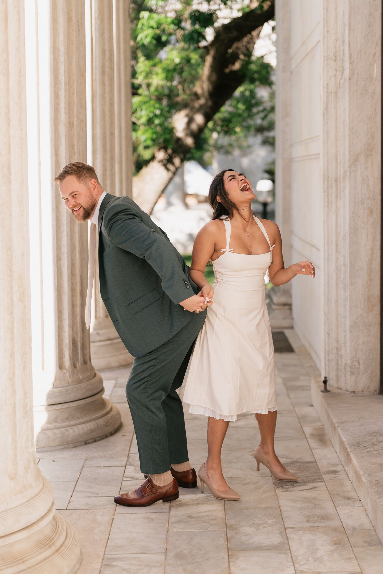 A laughing bride in a white dress and groom in a green suit hold hands playfully between marble columns.