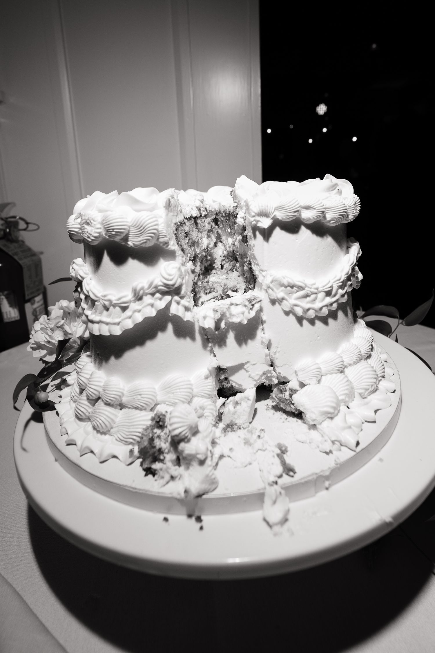 A black and white photo of a partially destroyed wedding cake with white frosting and decorative piping details on a plate.