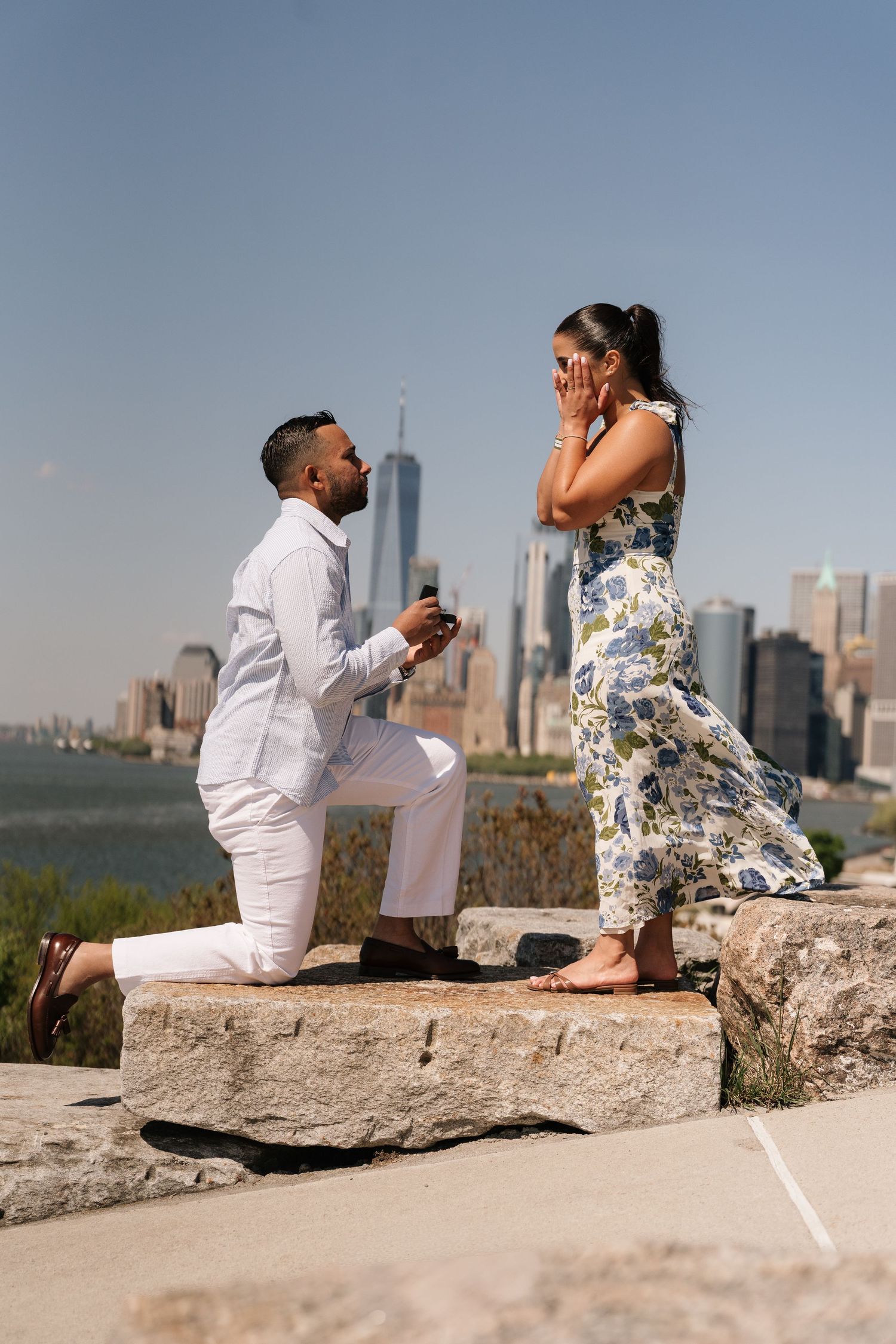 Man proposes to woman on one knee with NYC skyline backdrop on a sunny day.