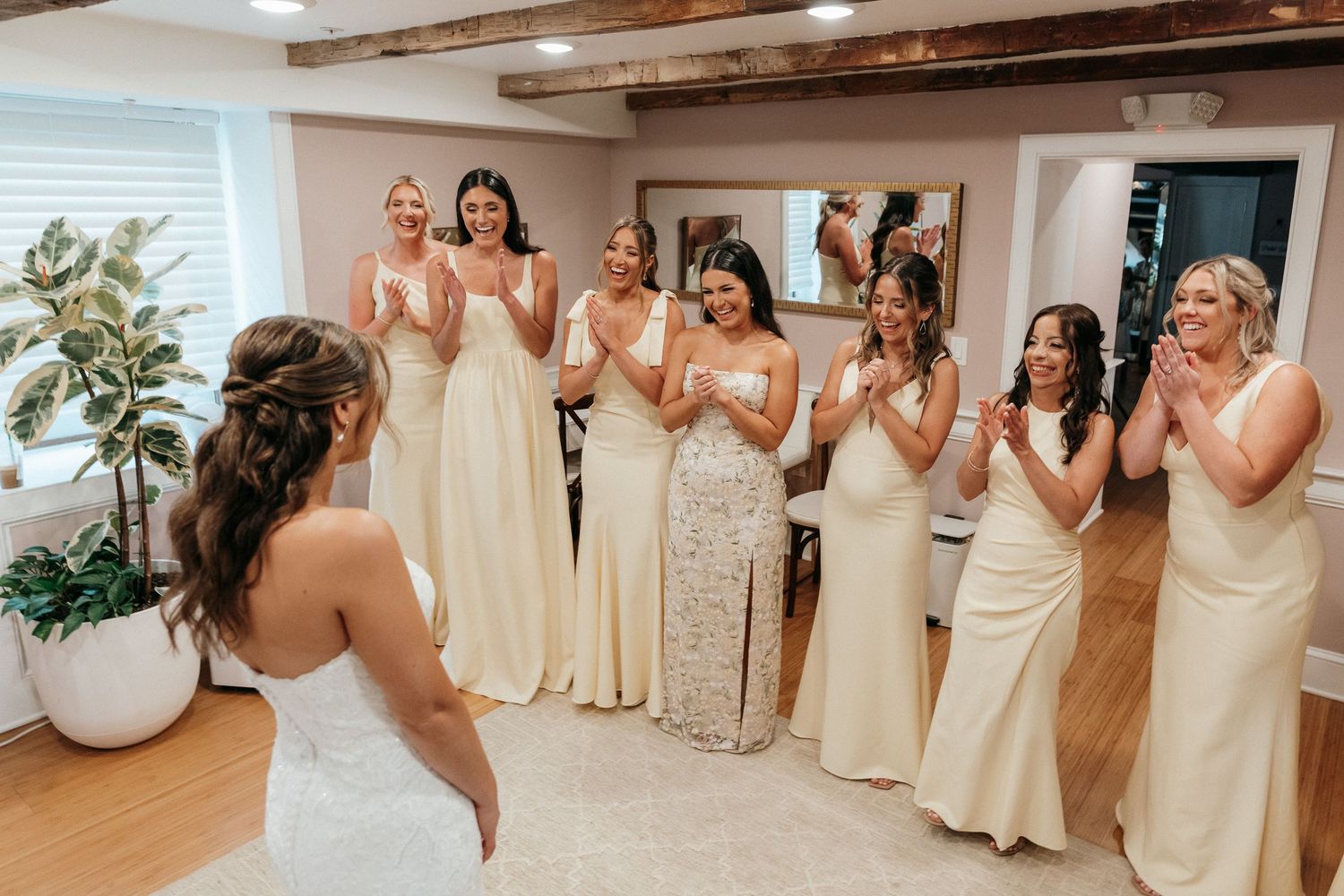 Bride reveals wedding dress to cheering bridesmaids in cream gowns during bridal party getting ready moment.