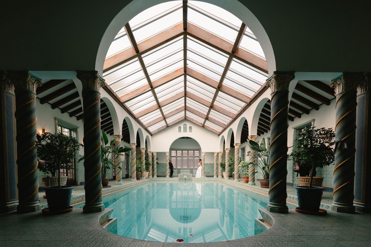 Interior view of an indoor pool with arched glass ceiling and ornate architectural columns.