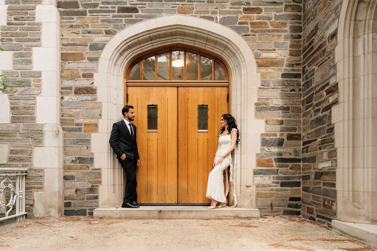 Couple posing in front of ornate wooden doors of a stone building, man in suit and woman in white dress.