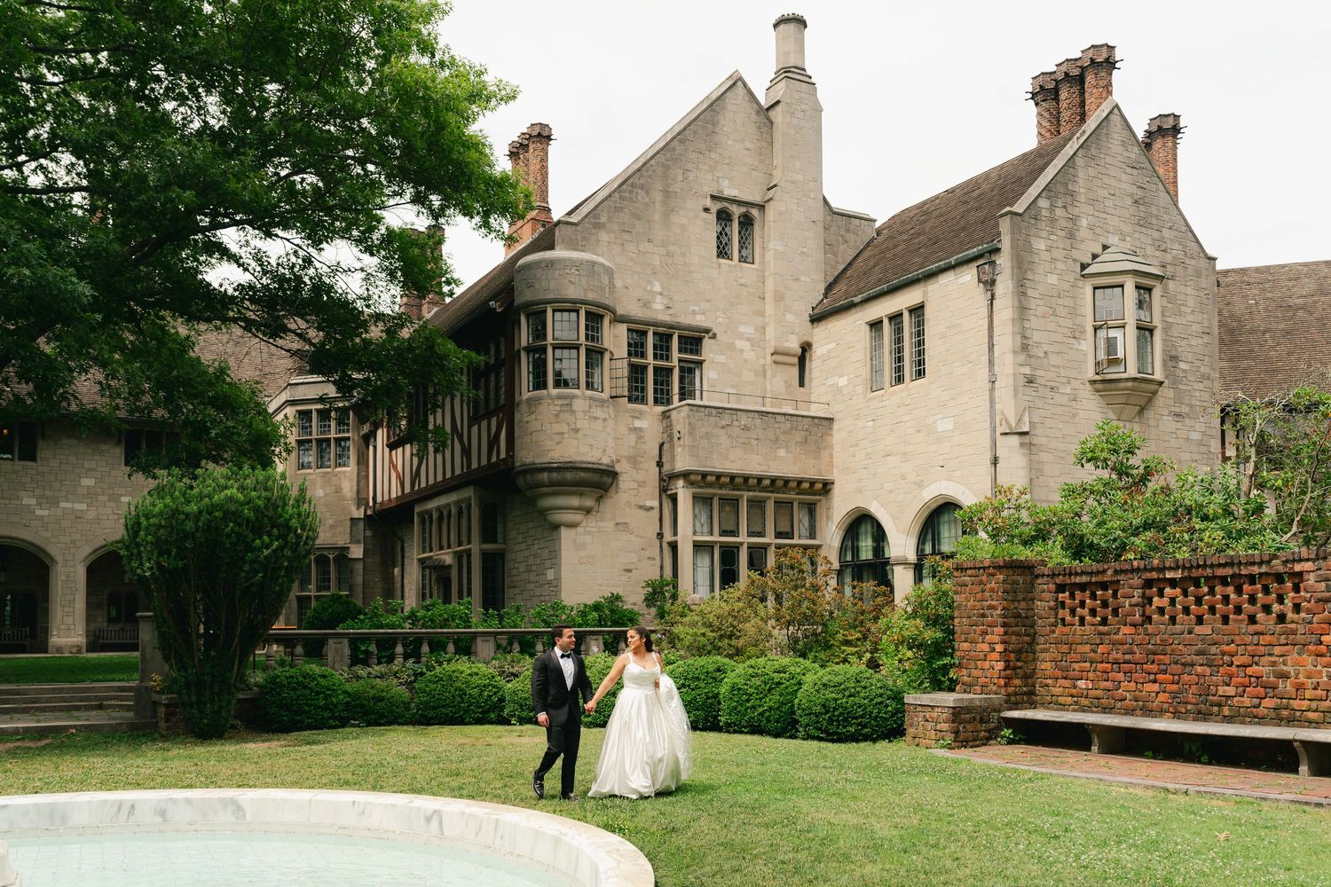 Bride and groom walk hand in hand on lush lawn outside elegant stone manor house wedding venue.