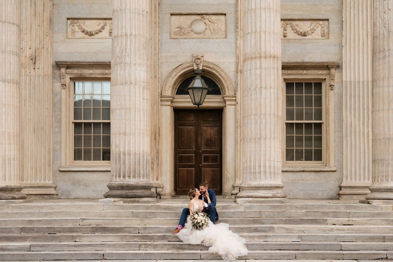 Bride and groom sitting on grand stone steps of a classical building with tall columns and ornate wooden doors.
