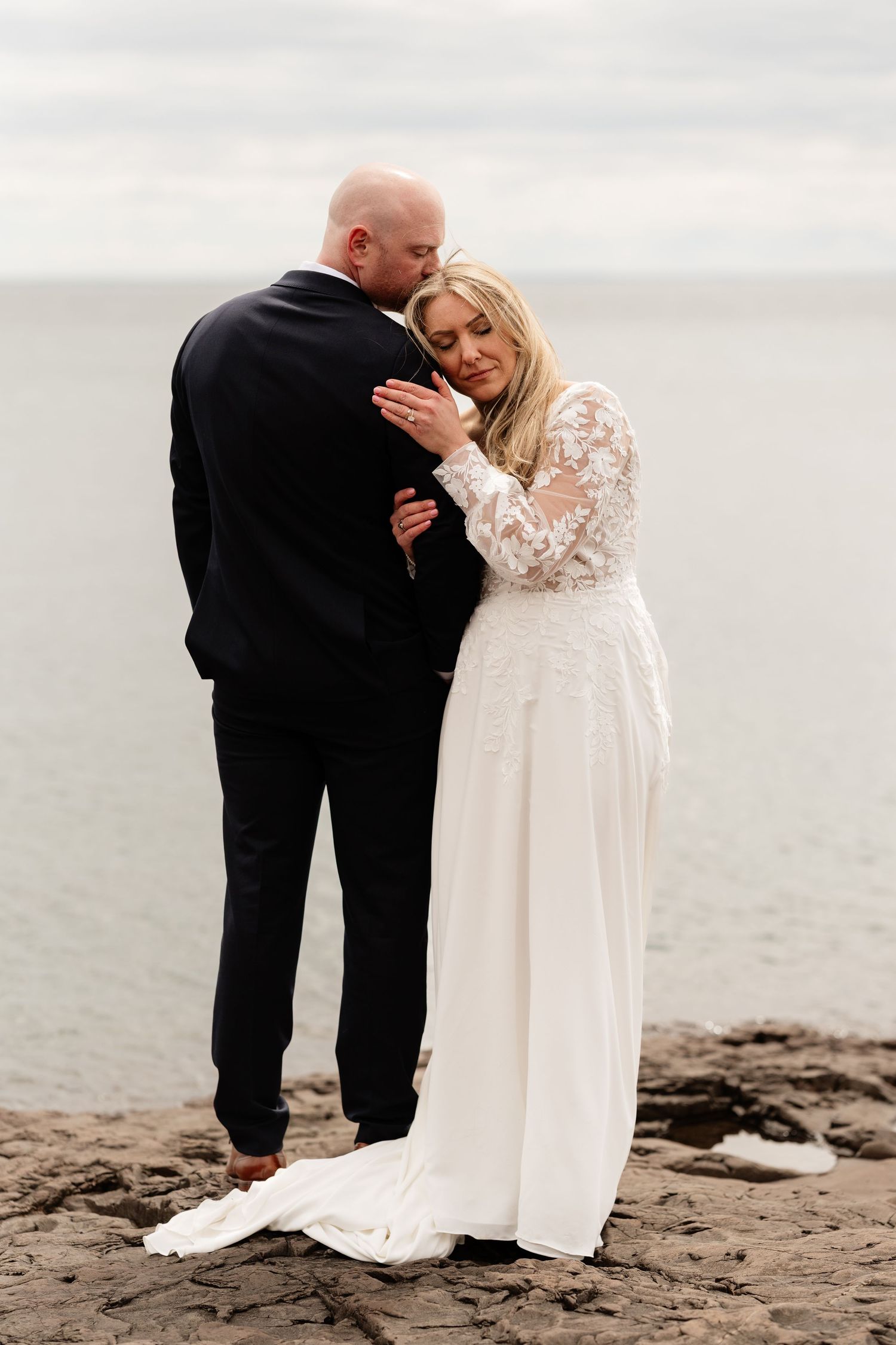 Bride in lace gown embraces groom in black suit on rocky shoreline with overcast sky backdrop.