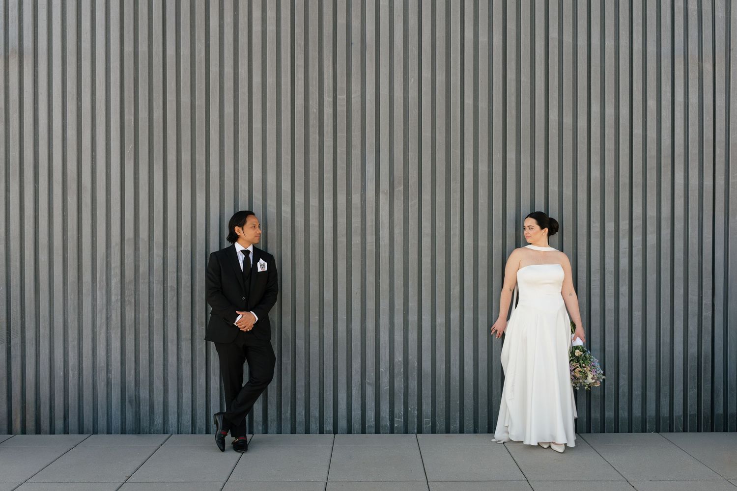 Bride in white gown and groom in black tuxedo stand apart against a modern corrugated metal wall.