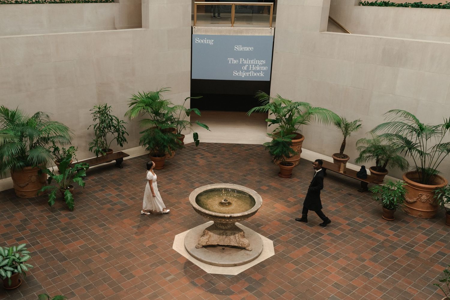 An aerial view of a museum atrium with a fountain, tropical plants, and two visitors walking on brick flooring.