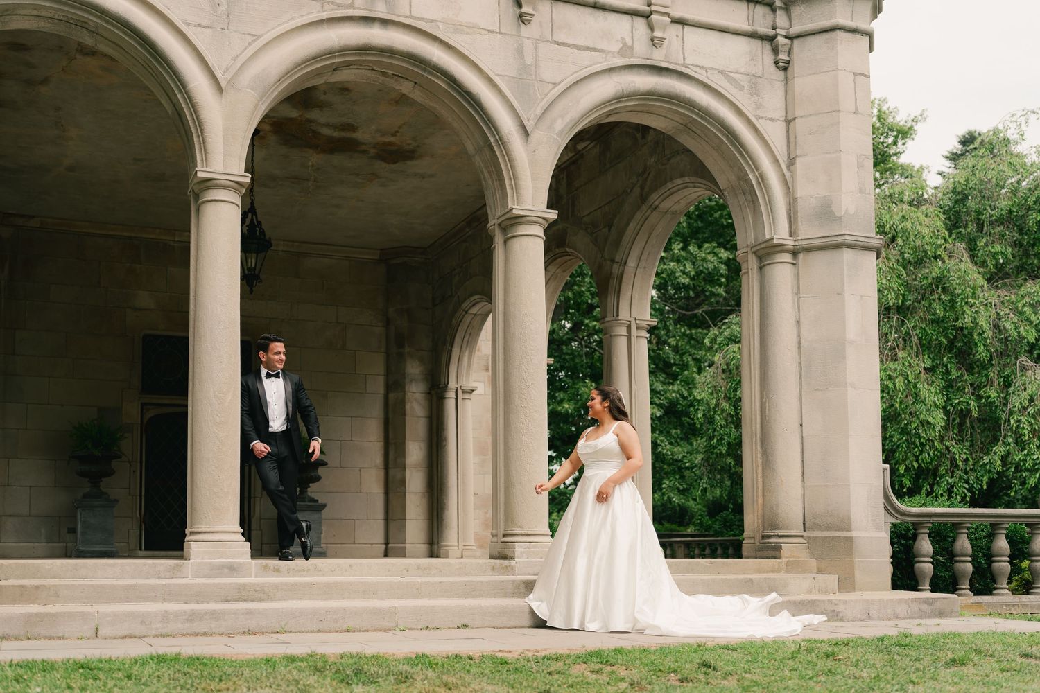 Bride and groom pose at elegant stone arched colonnade during outdoor wedding ceremony.