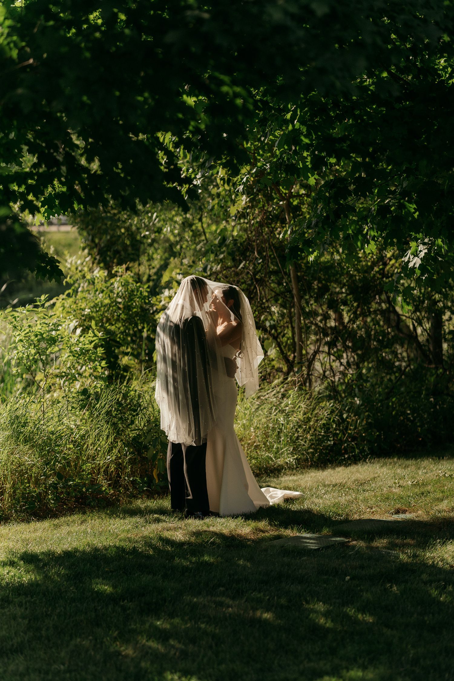 Bride and groom sharing a kiss under a wedding veil in a lush green garden setting.