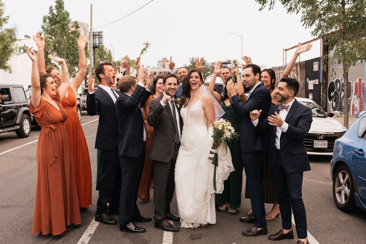 Wedding party celebrating with bride and groom on a city street, guests cheering in bridesmaid dresses and suits.