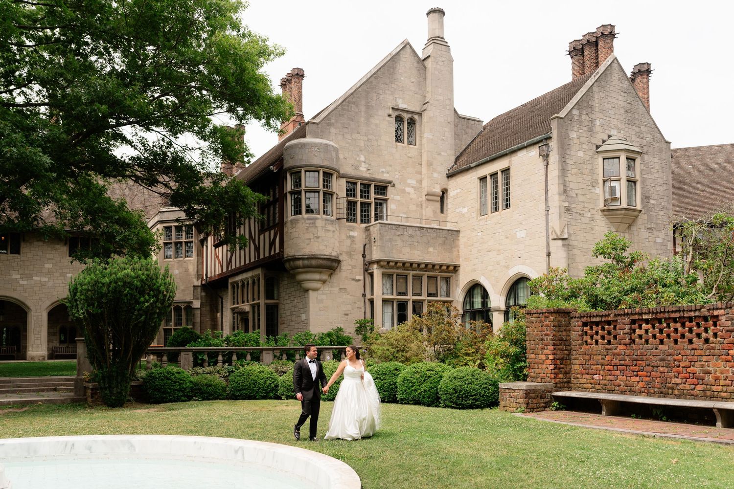 Bride and groom walk hand in hand on lush lawn outside elegant stone manor wedding venue.