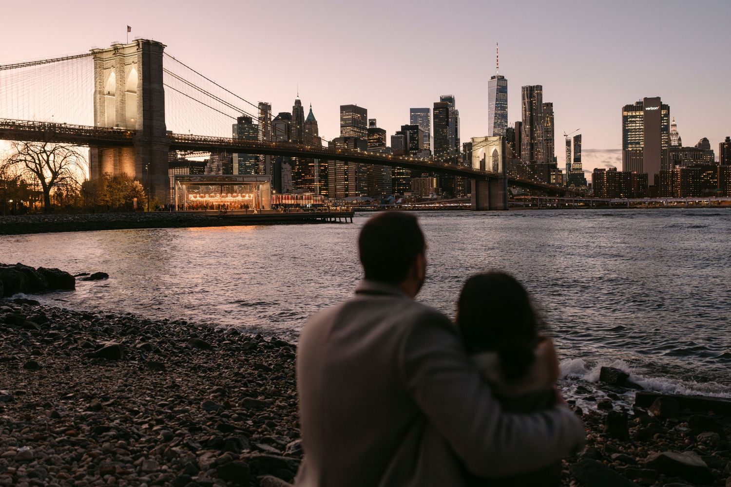 Romantic couple sits on rocky beach watching sunset over Manhattan skyline and East River from Brooklyn.