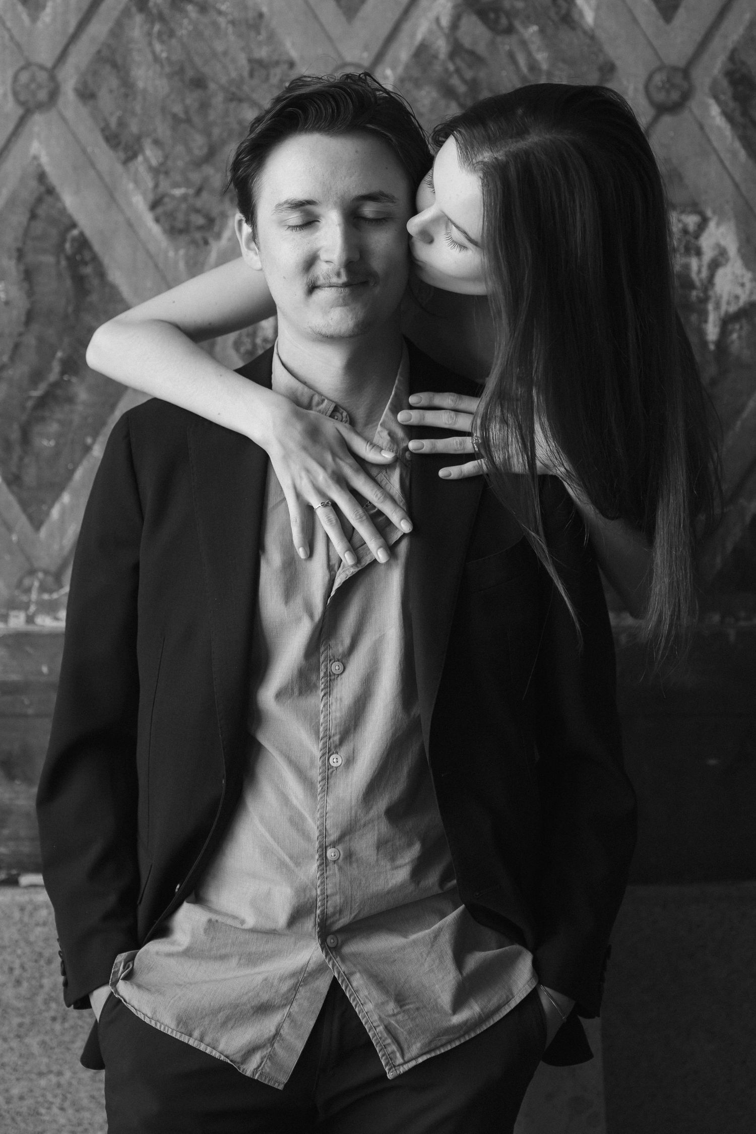 Black and white photo of a couple embracing, woman kissing mans cheek while he smiles with eyes closed.