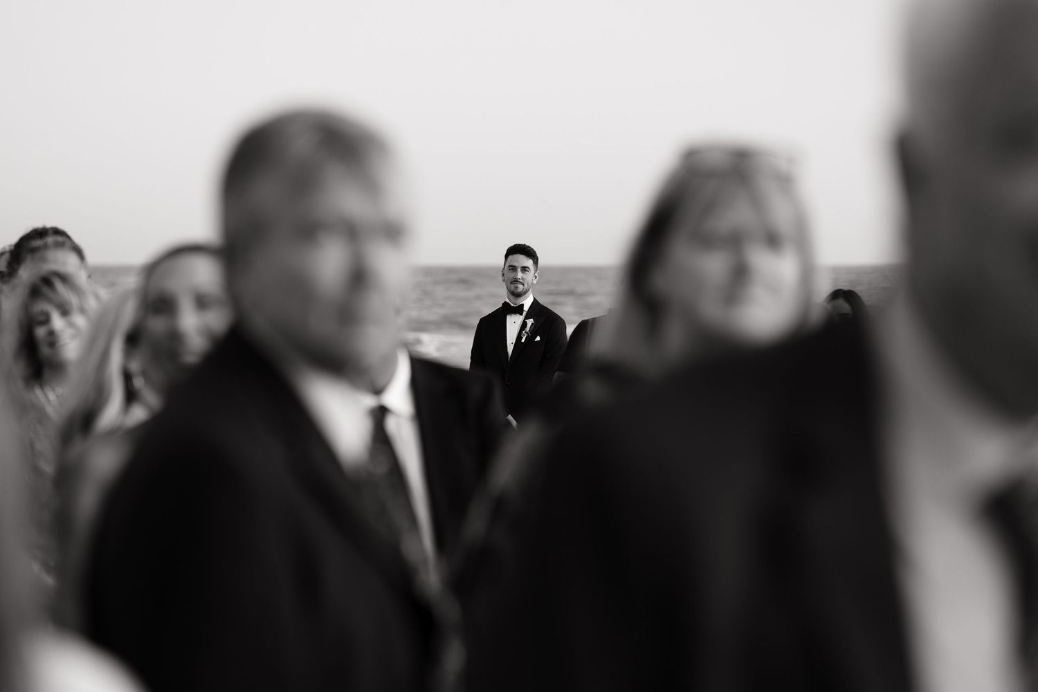 Creative black and white wedding photo with groom in focus while guests appear blurred in foreground.