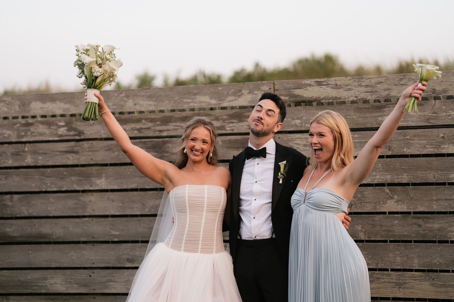 Wedding party members raise their arms in celebration while posing against a rustic wooden wall backdrop.