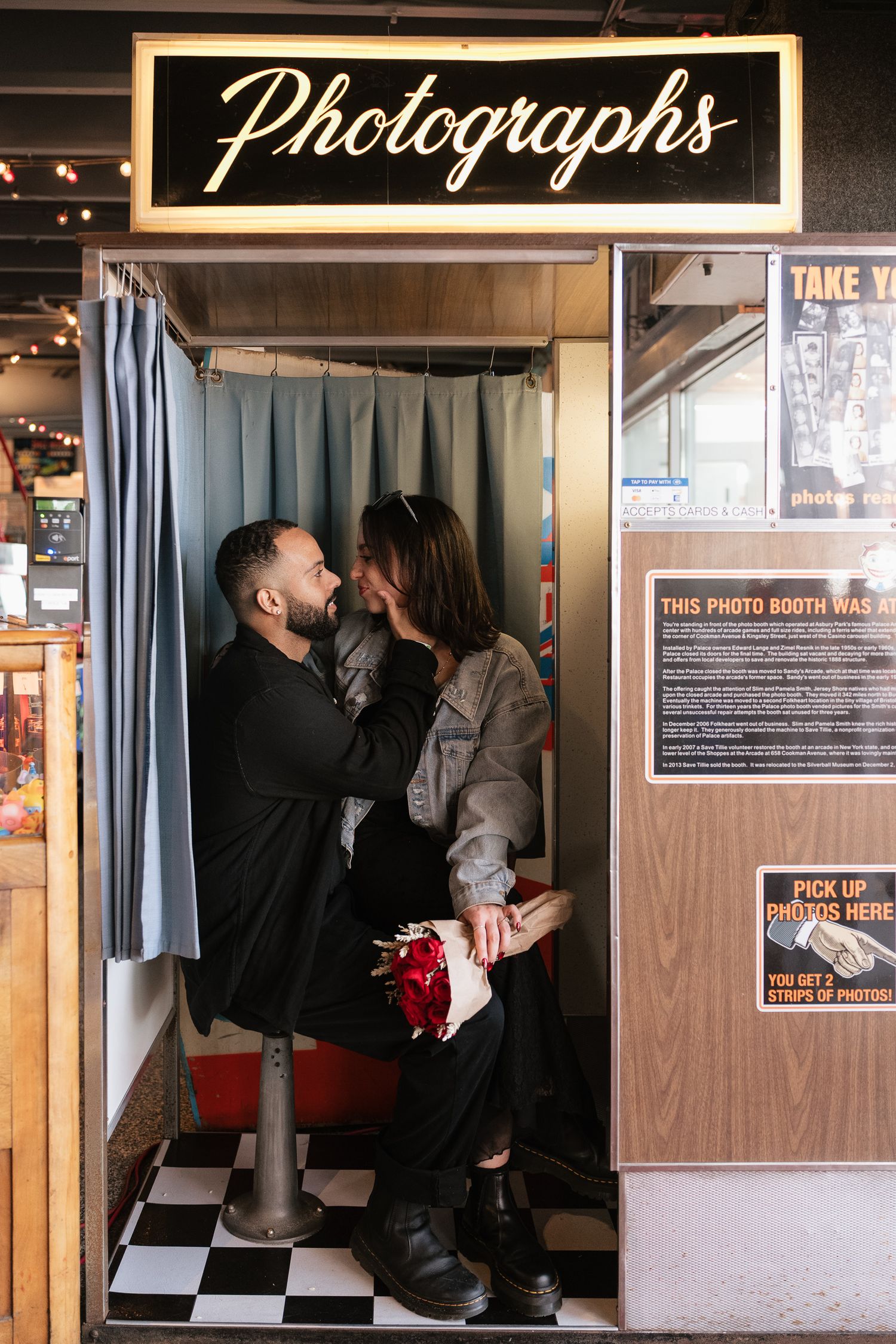 Couple sharing a romantic moment inside a vintage photo booth with a glowing Photographs sign above.