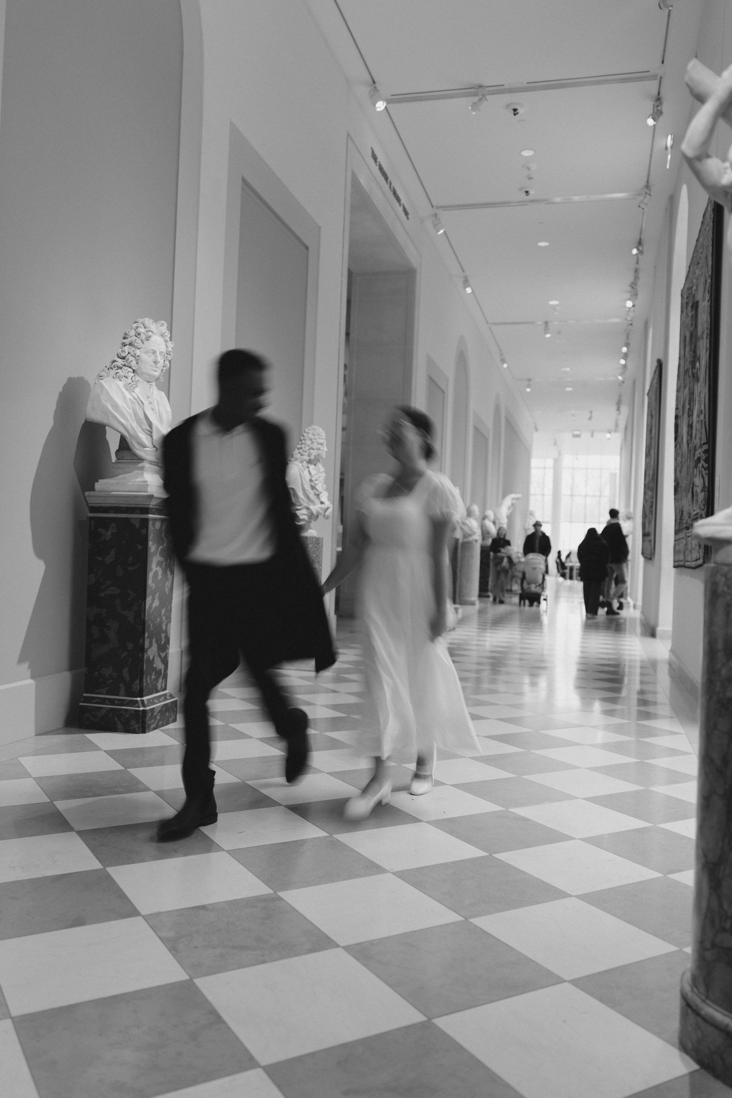 Motion-blurred figures walking through bright museum hallway with checkered floor.