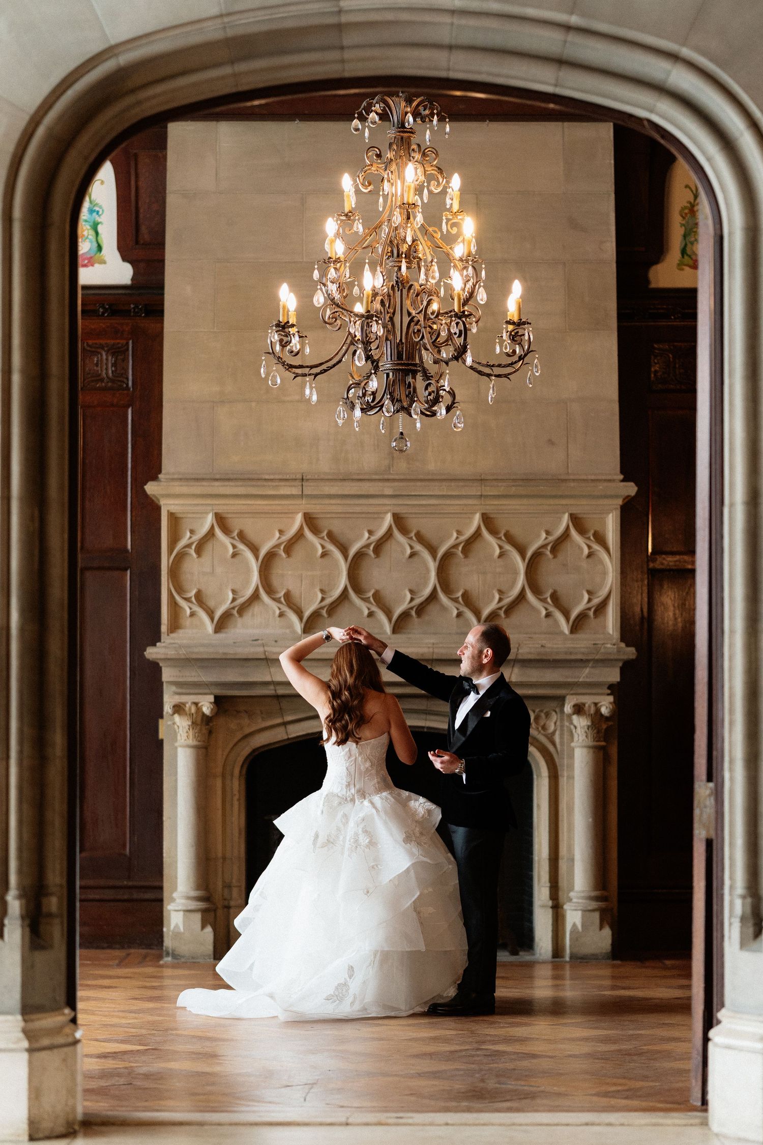 Bride and groom dancing under crystal chandelier by ornate fireplace in elegant ballroom.