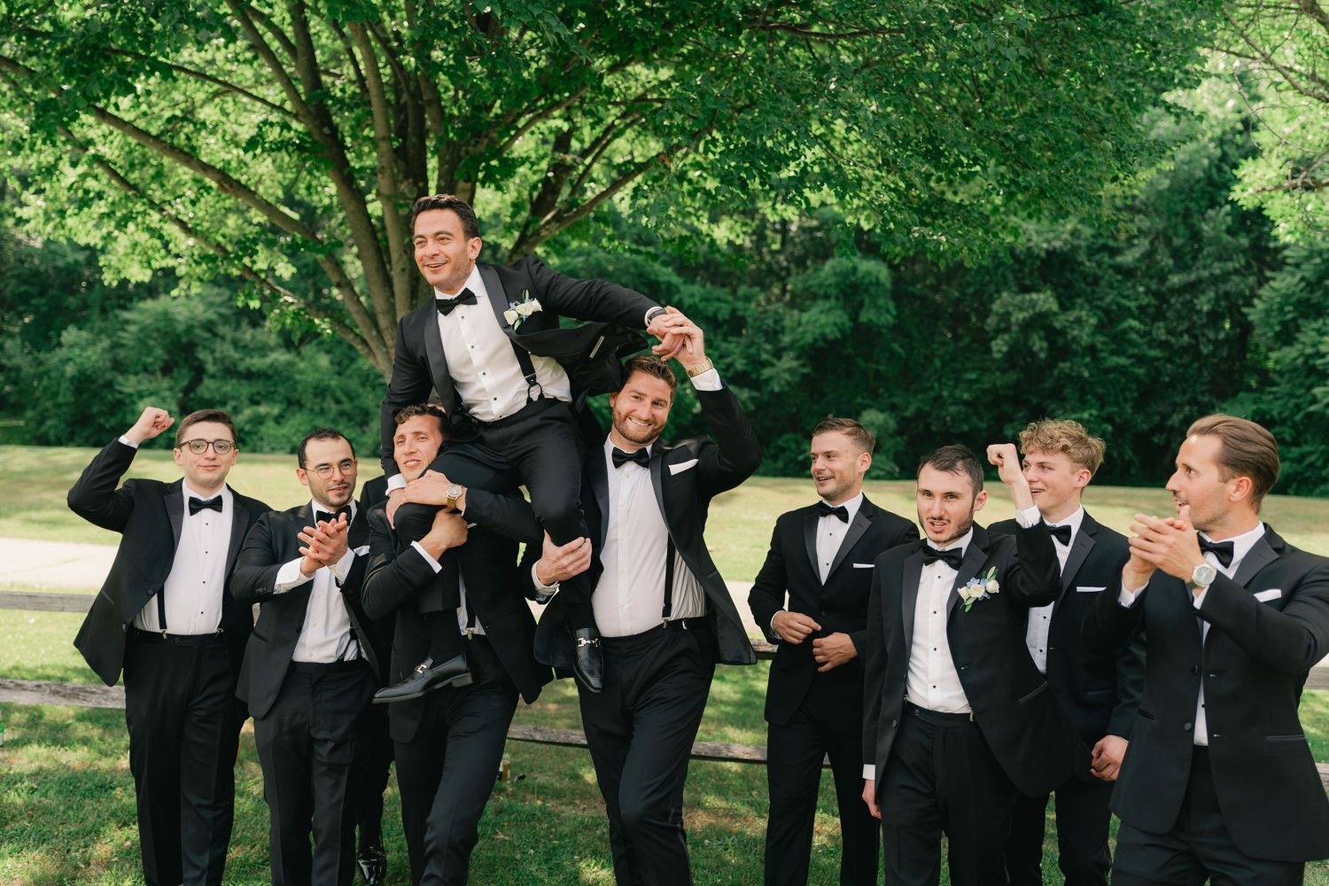 Groomsmen in black tuxedos lifting the groom outdoors near a tree at a wedding celebration.