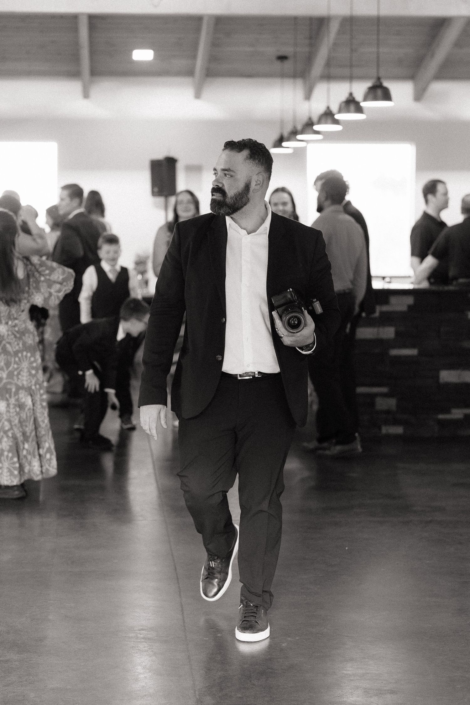 Black and white photo of a bearded man in a suit holding a camera walking through an elegant event venue.