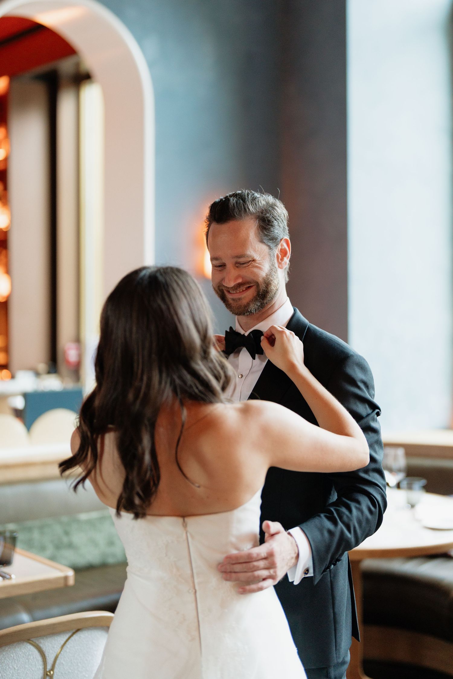 Smiling groom in black tuxedo adjusts bow tie as bride in strapless white gown faces him in elegant venue.