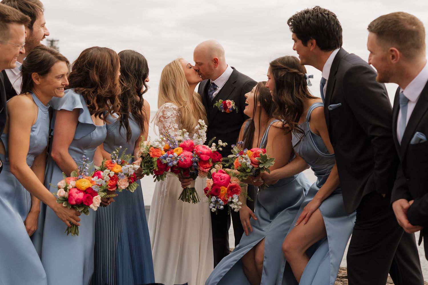 Wedding party kiss photo with bridesmaids in blue dresses holding colorful bouquets surrounding couple.