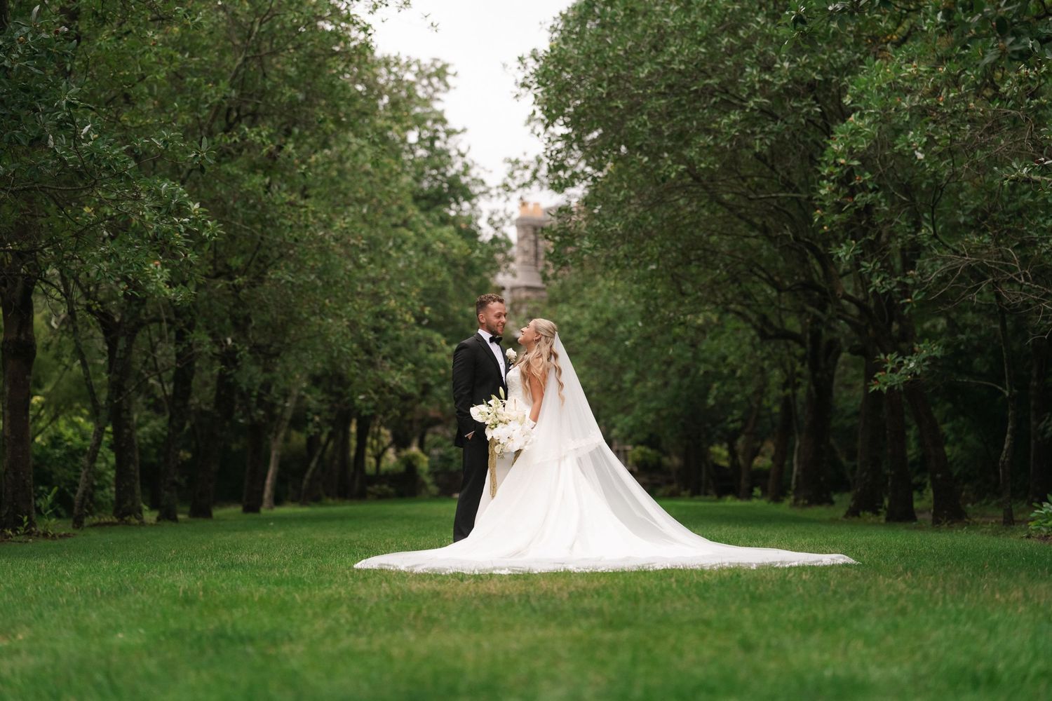 A bride and groom stand together in a lush green park, her flowing white gown and veil spread across the grass.
