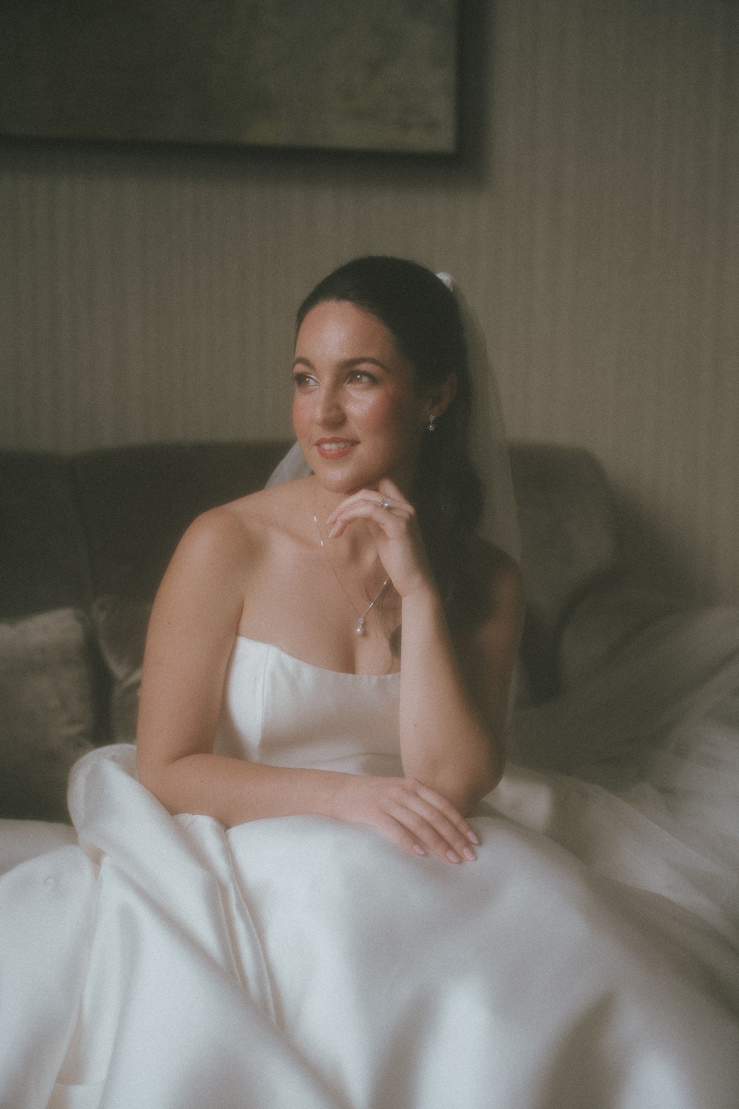 A bride in a white strapless gown sits gracefully on a sofa, gazing softly with a veil and delicate jewelry.