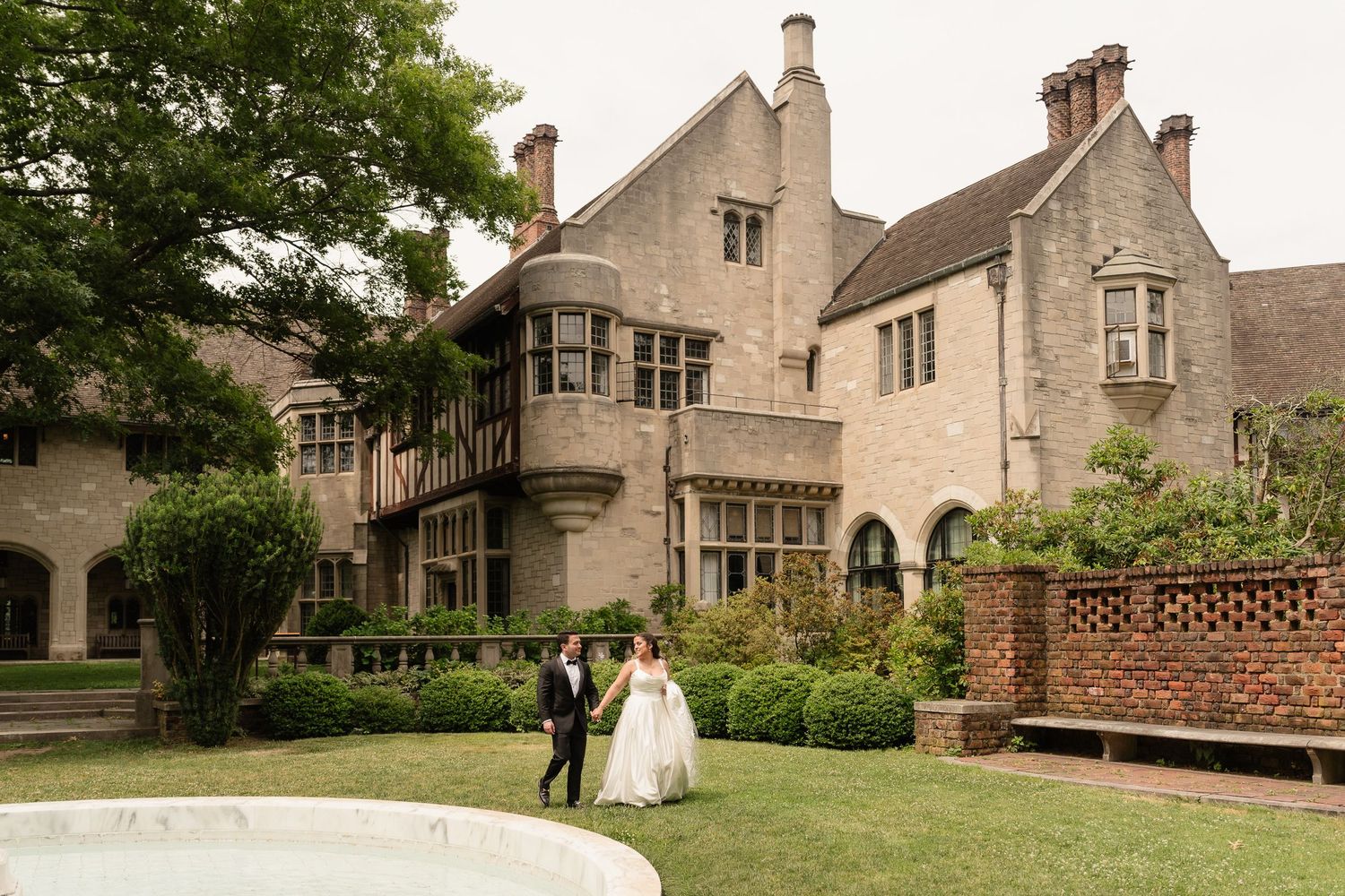 Bride and groom walk hand in hand on lush garden grounds of an elegant stone manor wedding venue.